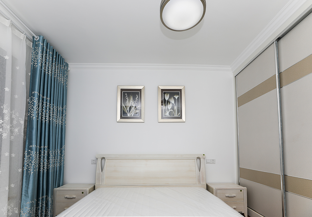 卧室是人们休息的主要场所，色调上以安静、深沉为主，蓝色的窗帘、白色的墙面这些安静色不仅能营造出安静、稳重的气氛，还能提高人的睡眠质量。黄白相间的大衣柜移门既稳重又温馨。
